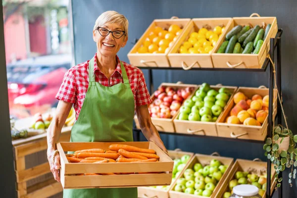 Mature woman works in fruits and vegetables shop. She is holding basket with carrot.