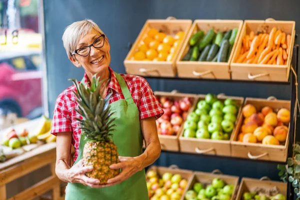 Mature woman works in fruits and vegetables shop. She is holding pineapple.
