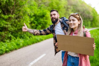 Yol kenarında otostop çeken mutlu çift arabayı durdurmaya çalışıyor. Mesajın için ellerinde boş karton var..