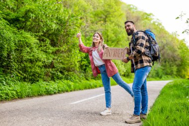 Yol kenarında otostop çeken mutlu çift arabayı durdurmaya çalışıyor. Üzerinde yazı olan bir karton tutuyorlar..