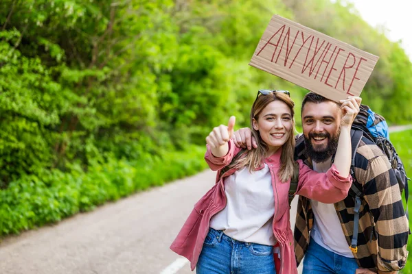 Happy couple hitchhiking on roadside trying to stop car. They are holding cardboard with inscription.