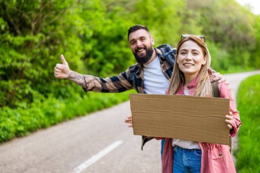 Yol kenarında otostop çeken mutlu çift arabayı durdurmaya çalışıyor. Mesajın için ellerinde boş karton var..