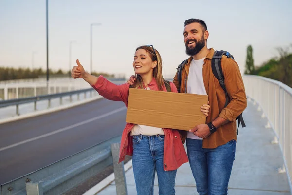 Happy couple hitchhiking on roadside trying to stop car. They are holding blank cardboard for your text.