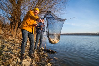 Father and son are fishing on sunny winter day. They caught a fish and are holding it in a landing net.