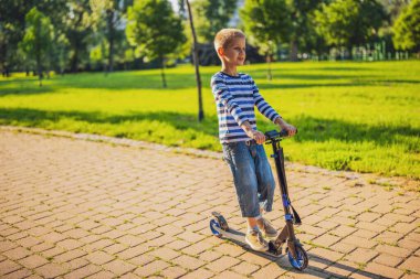 Çocuk güneşli bir günde parkta scooter sürüyor..