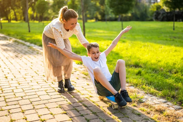 母は晴れた日に公園で息子と遊ぶ 男の子はスケートボードに乗っていると彼の母親は彼を押している — ストック写真
