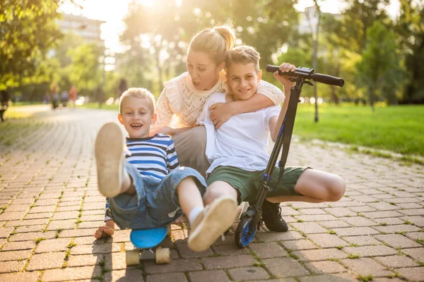 晴れた日に公園で彼女の2人の息子と楽しい母親 彼らはスケートボードやスクーターに乗っている — ストック写真