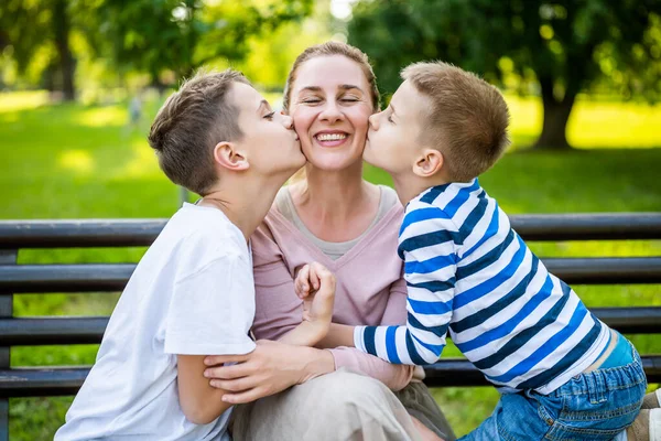 幸せな母親は公園で息子たちとベンチに座っている 彼らは一緒に楽しんでいる 男の子は母親とキスしてる — ストック写真
