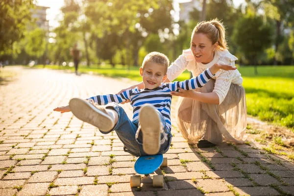 母は晴れた日に公園で息子と遊ぶ 男の子はスケートボードに乗っていると彼の母親は彼を押している — ストック写真