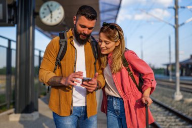 Mutlu çift tren istasyonunda duruyor ve trenlerinin gelmesini bekliyor. Akıllı telefondan haritaya bakıyorlar..
