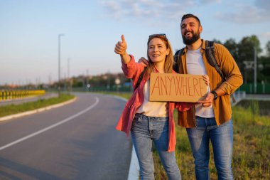 Yol kenarında otostop çeken mutlu çift arabayı durdurmaya çalışıyor. Üzerinde yazı olan bir karton tutuyorlar..