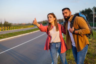 Yetişkin bir çift yol kenarında otostop çekerek arabayı durdurmaya çalışıyor. Endişeli ve sıkılmış durumdalar..