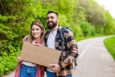 Yol kenarında otostop çeken mutlu çift arabayı durdurmaya çalışıyor. Mesajın için ellerinde boş karton var..