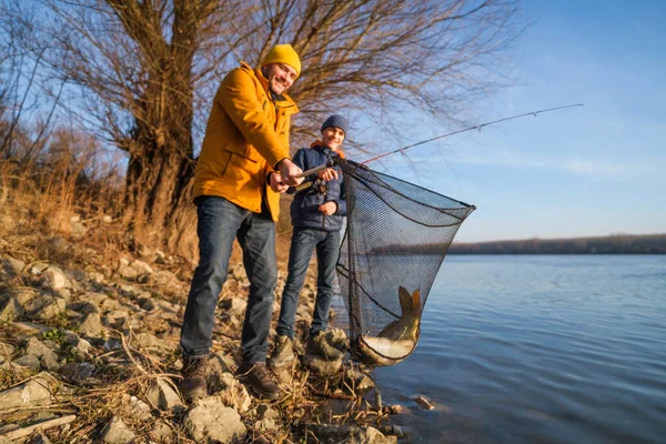 Father and son are fishing on sunny winter day. They caught a fish and are holding it in a landing net.