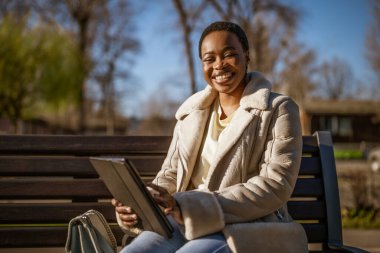 Güneşli bir günde mutlu bir Afro-Amerikan kadının açık hava portresi. Sokakta bankta oturuyor ve dijital tablet kullanıyor..