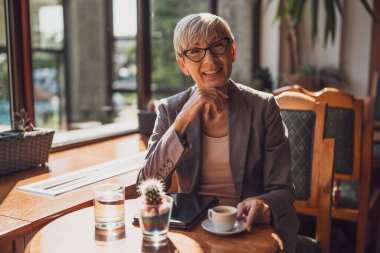 Olgun kadın kafede oturuyor ve dinleniyor. Kahve içiyor ve akıllı telefon kullanıyor..