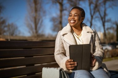 Güneşli bir günde mutlu bir Afro-Amerikan kadının açık hava portresi. Sokakta bankta oturuyor ve dijital tablet kullanıyor..