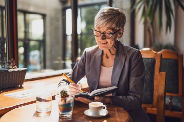 Olgun kadın kafede oturuyor ve dinleniyor. Kahve içiyor ve not defteri kullanıyor..
