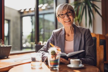 Olgun kadın kafede oturuyor ve dinleniyor. Kahve içiyor ve not defteri kullanıyor..