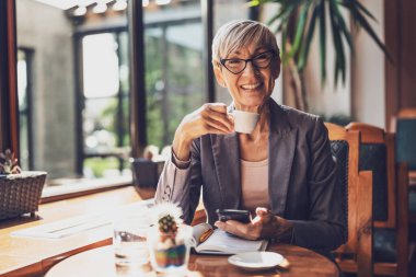 Olgun kadın kafede oturuyor ve dinleniyor. Kahve içiyor ve telefonda mesajlaşıyor..