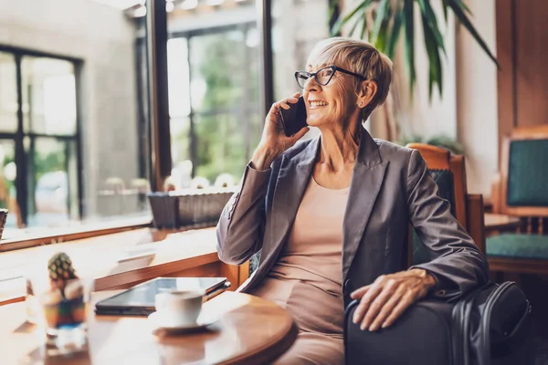 stock image Mature woman is sitting in cafe and relaxing. She is drinking coffee and talking on phone.