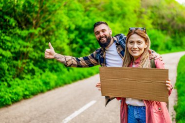 Yol kenarında otostop çeken mutlu çift arabayı durdurmaya çalışıyor. Mesajın için ellerinde boş karton var..