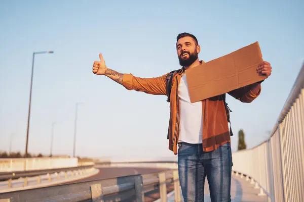Man is hitchhiking on roadside trying to stop car. He is holding blank cardboard for your text.