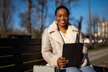 Güneşli bir günde mutlu bir Afro-Amerikan kadının açık hava portresi. Sokakta bankta oturuyor ve dijital tablet kullanıyor..
