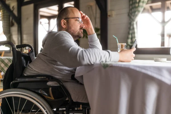 Paraplegic handicapped man in wheelchair is sitting at restaurant and using smartphone.