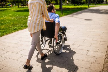 Tekerlekli sandalyedeki adam annesiyle parkta vakit geçiriyor. Birlikte güneşli bir günün tadını çıkarıyorlar..