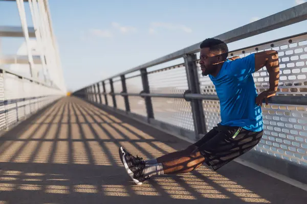 若いアフリカ系アメリカ人男性が市内の橋で運動している 彼は逆のプッシュアップをしている ロイヤリティフリーのストック写真