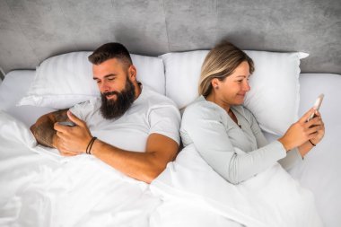 Genç mutlu çift sabahları yatakta yatıyor. Rahatlıyorlar ve akıllı telefonlar kullanıyorlar..