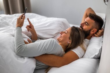 Genç ve mutlu bir çift sabahları yatakta yatıyor. Rahatlıyorlar ve akıllı telefon kullanıyorlar..