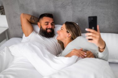 Genç ve mutlu bir çift sabahları yatakta yatıyor. Rahatlıyorlar ve akıllı telefon kullanıyorlar..