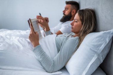 Genç çift akşamları yatakta yatıyor. Kadın kitap okuyor ve erkek akıllı telefon kullanıyor..