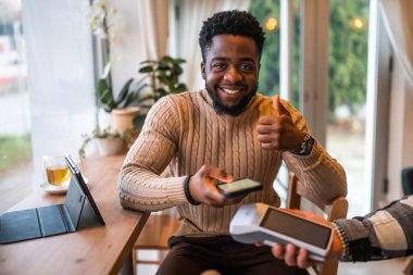 Rahat bir kafede otururken, günlük giysiler içindeki genç adam akıllı telefonuyla temas kurmadan ödeme yapıyor..