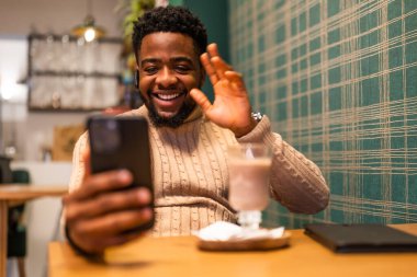 Neşeli genç adam internette sohbet ediyor. Akıllı telefon kullanıyor, video görüşmesi yapıyor ve sıcak çikolata içiyor..