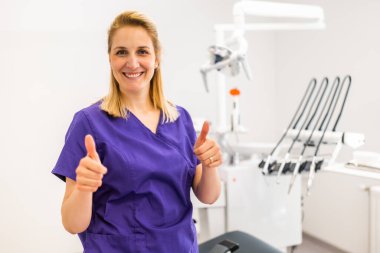 Dişçinin muayenehanesinde mutlu bir dişçinin portresi. Dişçi kliniğinde çalışan kendine güvenen yetişkin bir kadın. Başparmağını kaldırıyor..