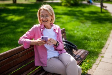 Parktaki mutlu olgun kadın portresi. Bankta oturuyor ve akıllı telefon kullanıyor..