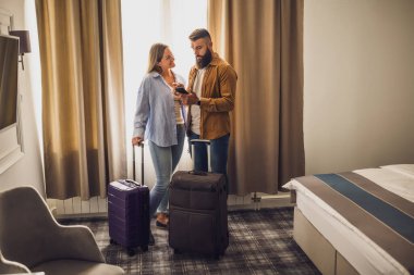 30 'lu yaşlarda bir çift otel odasında bavulları açık bekliyor. Akıllı telefon kullanıyorlar ve kalacakları yeri planlıyorlar..