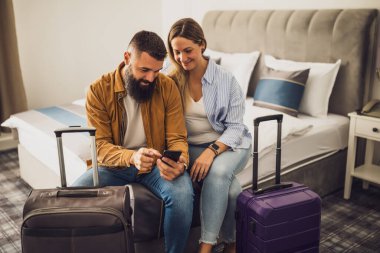 Mutlu evli çift otel odasında yatakta oturuyor ve akıllı telefon kullanarak tatillerini planlıyorlar. Bavullar yatağın yanında..