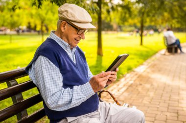 Şapkalı yaşlı adam güneşli bir günde parkta bankta oturuyor ve dijital tablet kullanıyor..