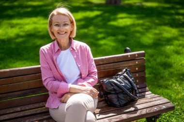 Mutlu olgun kadın güneşli bir günde parkta bankta oturuyor. Kameraya bakıyor ve gülümsüyor..