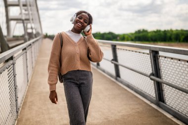 Mutlu genç siyah kadın köprü yolunda yürüyor, müzik dinliyor ve gülümsüyor. Dışarıda güneşli bir günün tadını çıkarıyor..