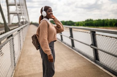 Mutlu genç siyah kadın köprü yolunda yürüyor, müzik dinliyor ve gülümsüyor. Dışarıda güneşli bir günün tadını çıkarıyor..