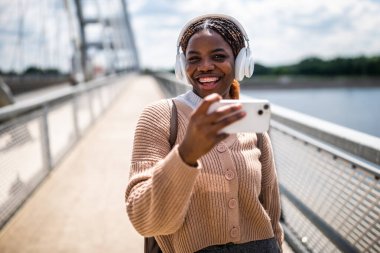Mutlu genç siyah kadın kameraya bakıyor ve gülümsüyor. Açık havada güneşli bir günün tadını çıkarıyor ve fotoğraf çekerken müzik dinliyor..