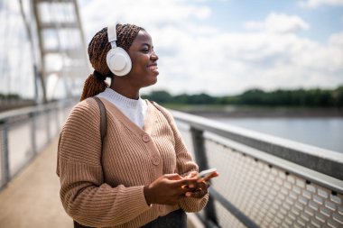 Mutlu genç siyah kadın kameraya bakıyor ve gülümsüyor. Açık havada güneşli bir günün tadını çıkarıyor ve akıllı telefon kullanarak müzik dinliyor..