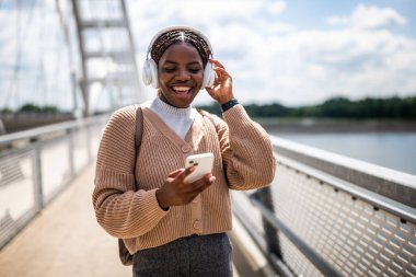 Mutlu genç siyah kadın kameraya bakıyor ve gülümsüyor. Açık havada güneşli bir günün tadını çıkarıyor ve akıllı telefon kullanarak müzik dinliyor..