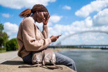 Genç endişeli siyah kadın akıllı telefon kullanıyor. Şehirdeki nehrin kenarında oturuyor..
