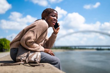 Genç, neşeli siyahi kadın telefonda konuşuyor. Gülüyor ve şehirde güneşli bir günün tadını çıkarıyor..
